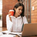 brunette-woman-drinking-coffee-to-go-while-working-on-laptop-e1703388822223.jpg