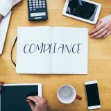 compliance-headline-on-paper-notebook-at-small-business-office-desk-with-young-adult-workers-e1706684795841.jpg